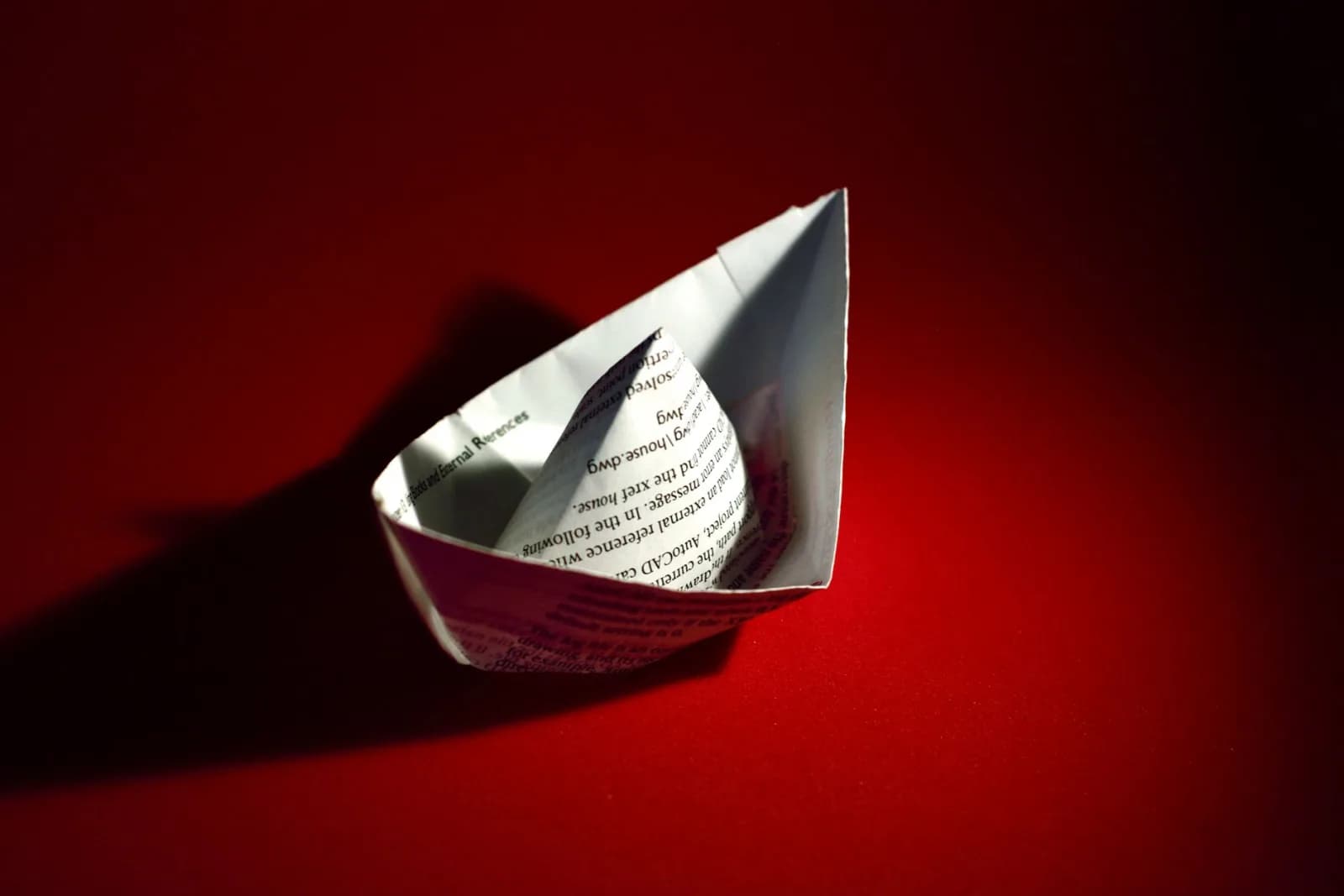 A paper boat crafted from newspaper, set against a vivid red background with shadows and highlights.