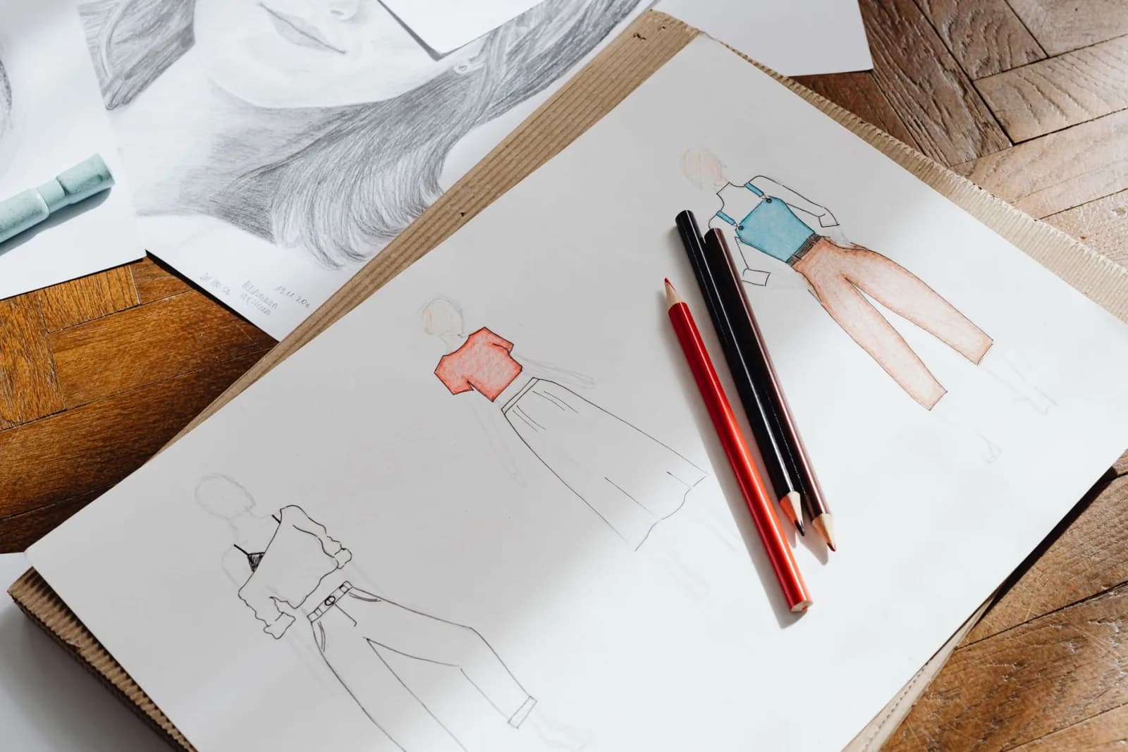 Artistic fashion design sketches on paper accompanied by colored pencils on a wooden surface.