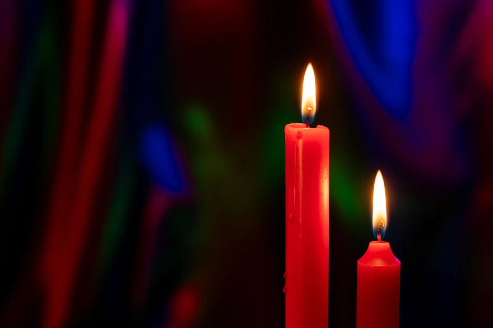 Two red candles with vibrant flames stand out against a colorful bokeh background.