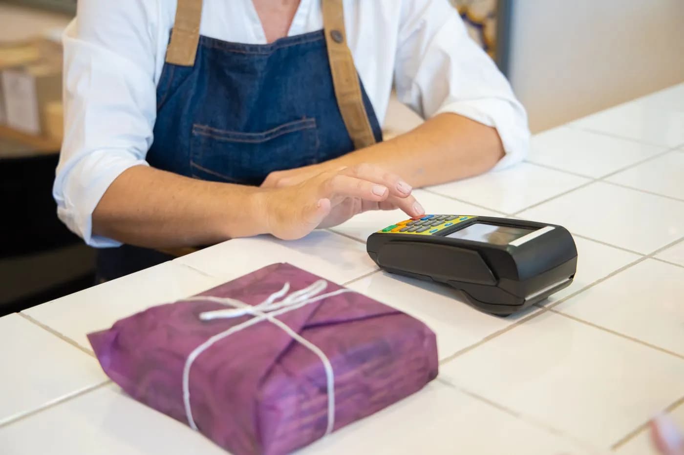 A shopkeeper at the counter with a card terminal beside a wrapped parcel