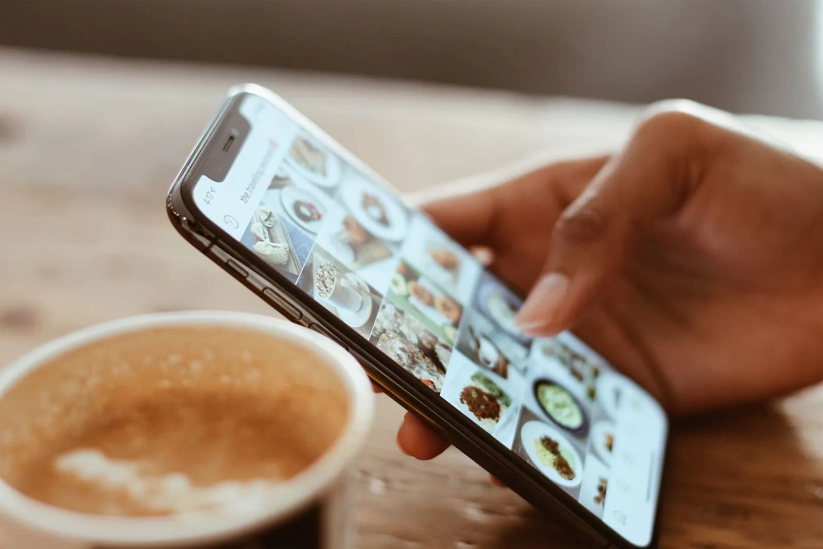 A hand scrolling social media on a phone beside a coffee