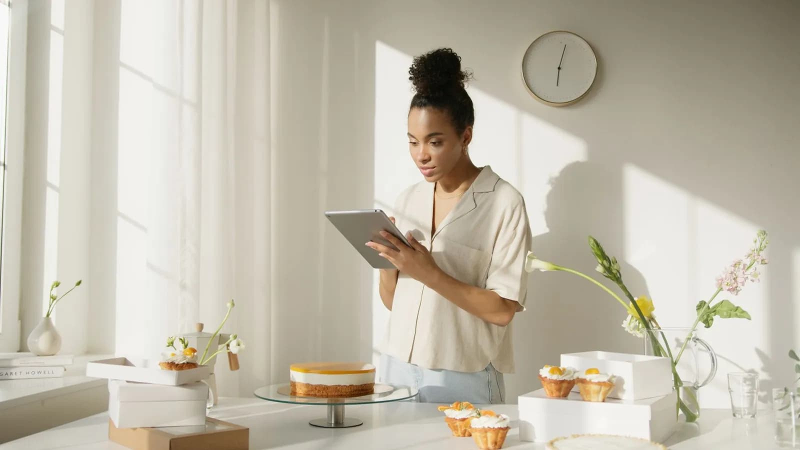 Une femme utilisant une tablette dans une cuisine lumineuse avec des gâteaux et des fleurs