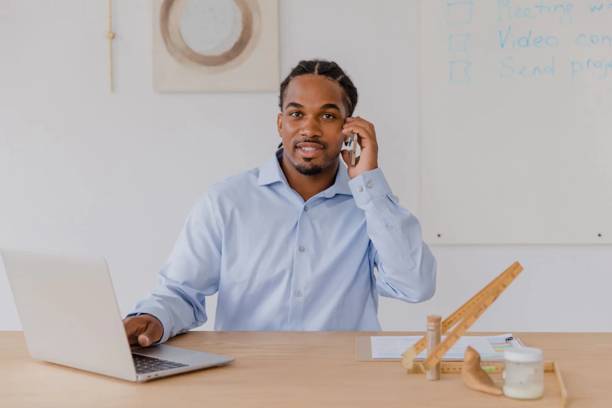 A young man at his office desk on the phone, smiling at his laptop