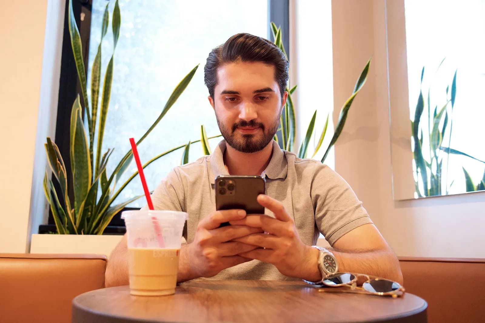 A young man checking his phone in a cafe with plants and an iced drink