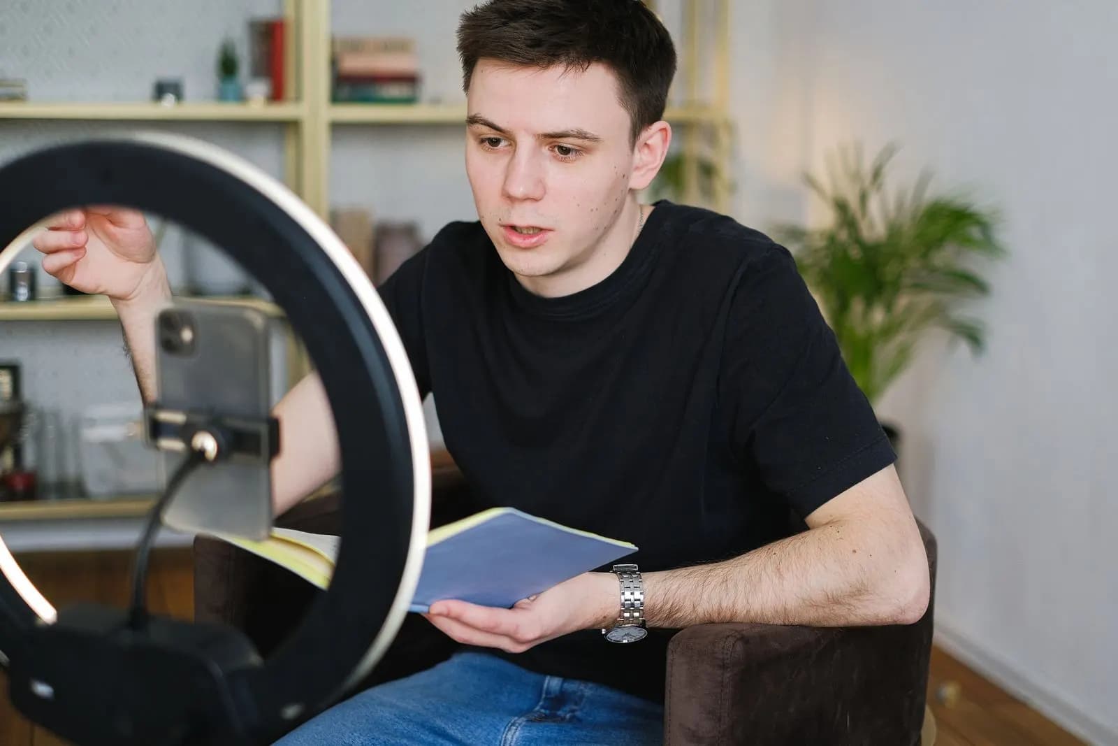 A man recording a video with a phone and a ring light, holding a notebook
