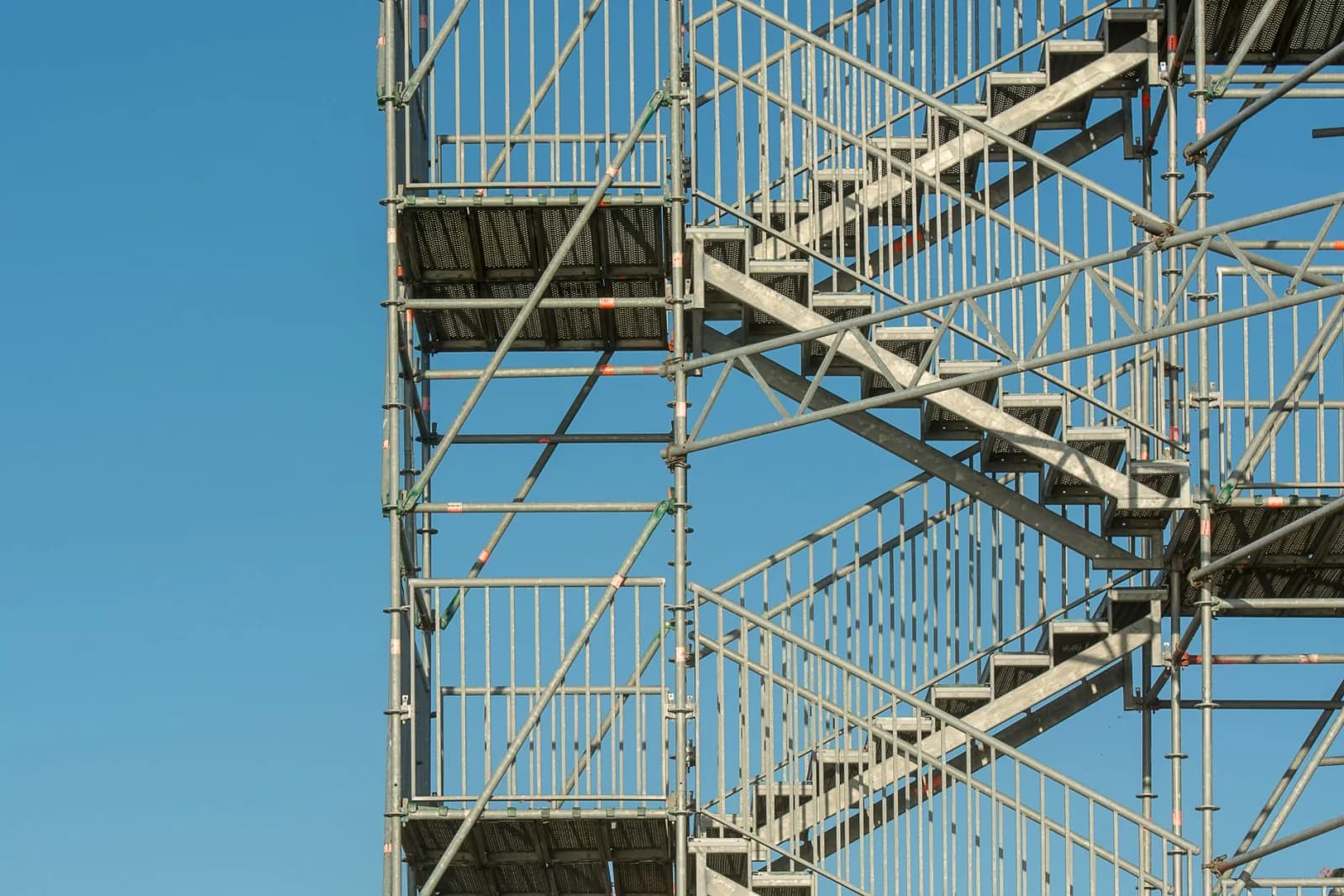 Close-up of metal scaffolding structure with stairs against a clear blue sky, illustrating construction and industrial themes.