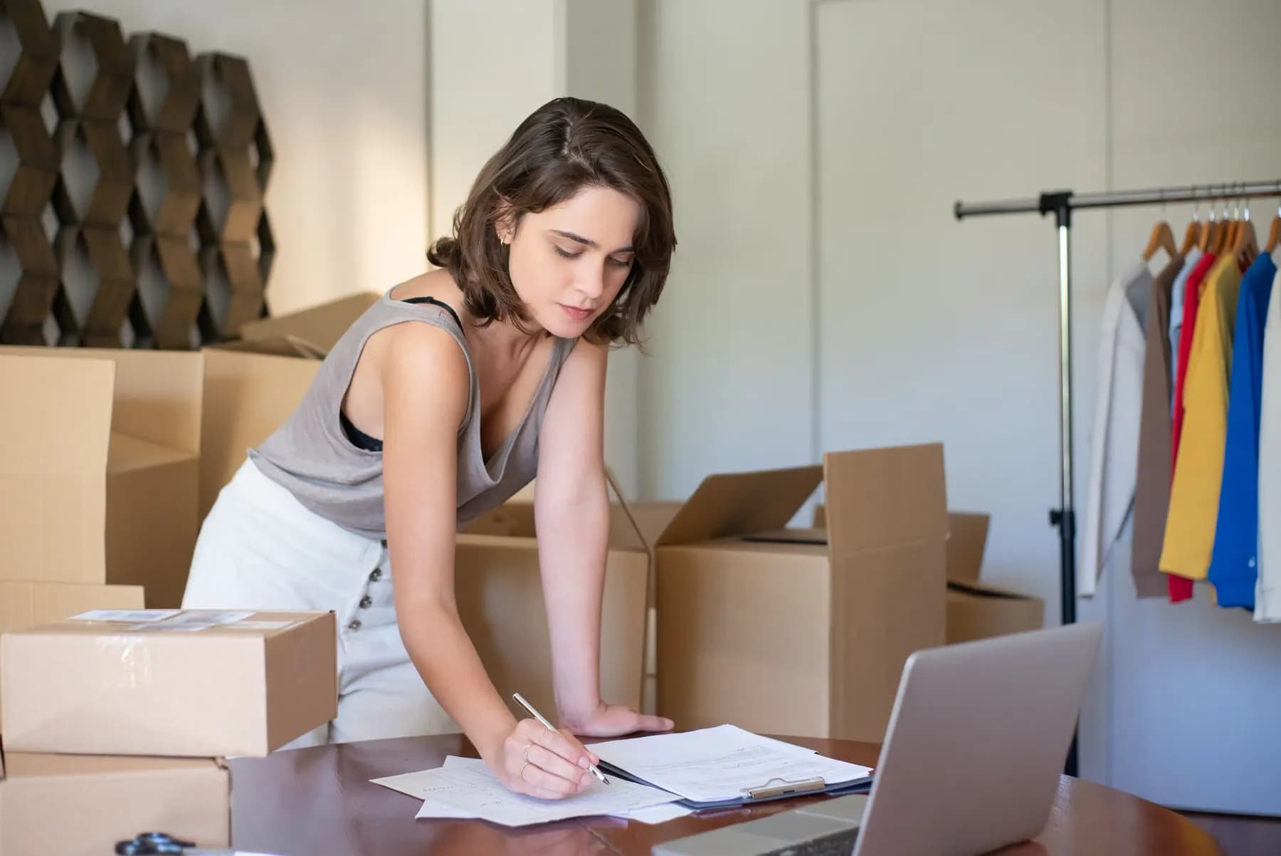 An online clothing seller managing orders on a laptop between parcels and a clothing rail