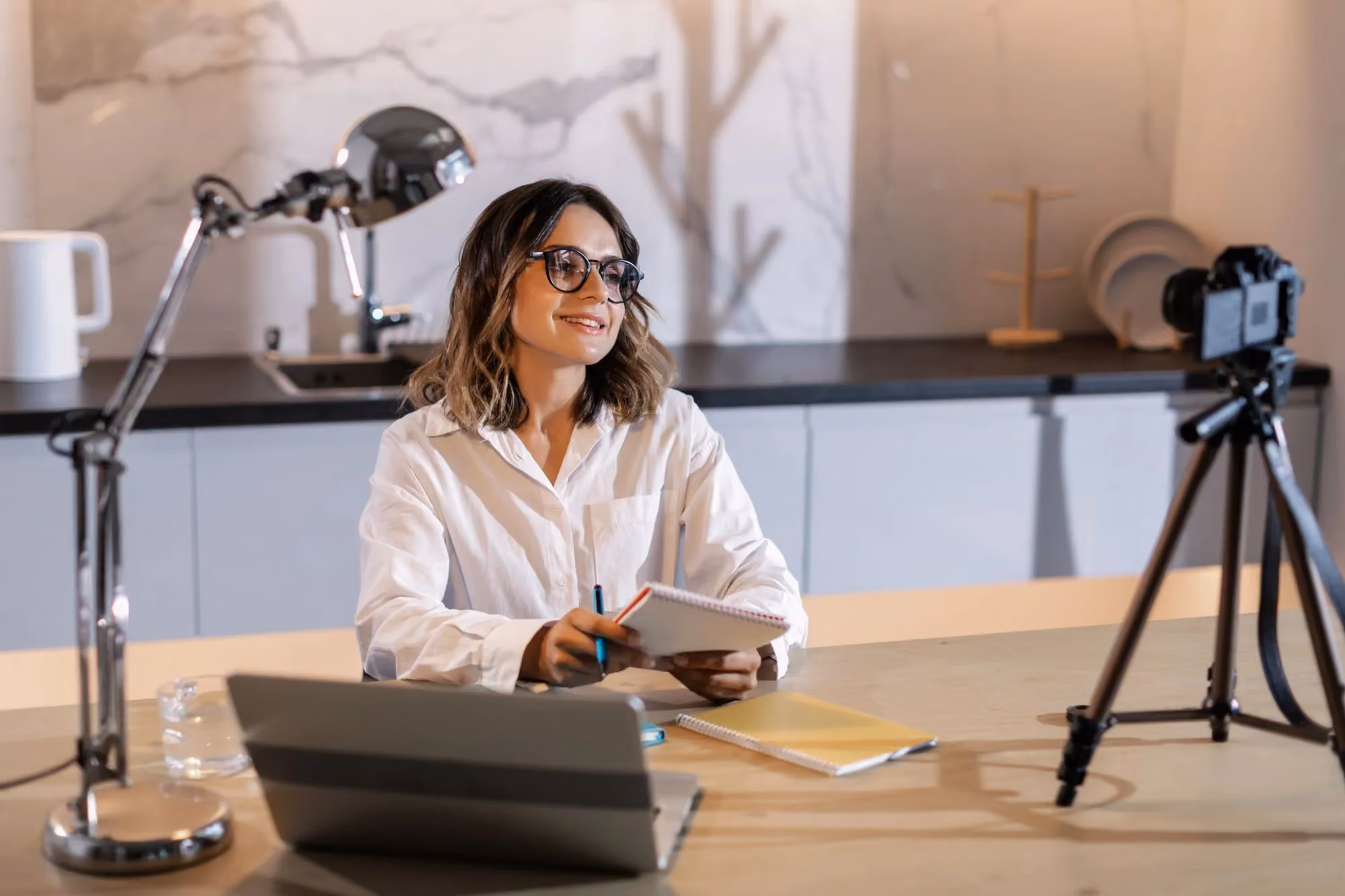 A smiling woman taking notes at her desk beside a laptop and a camera